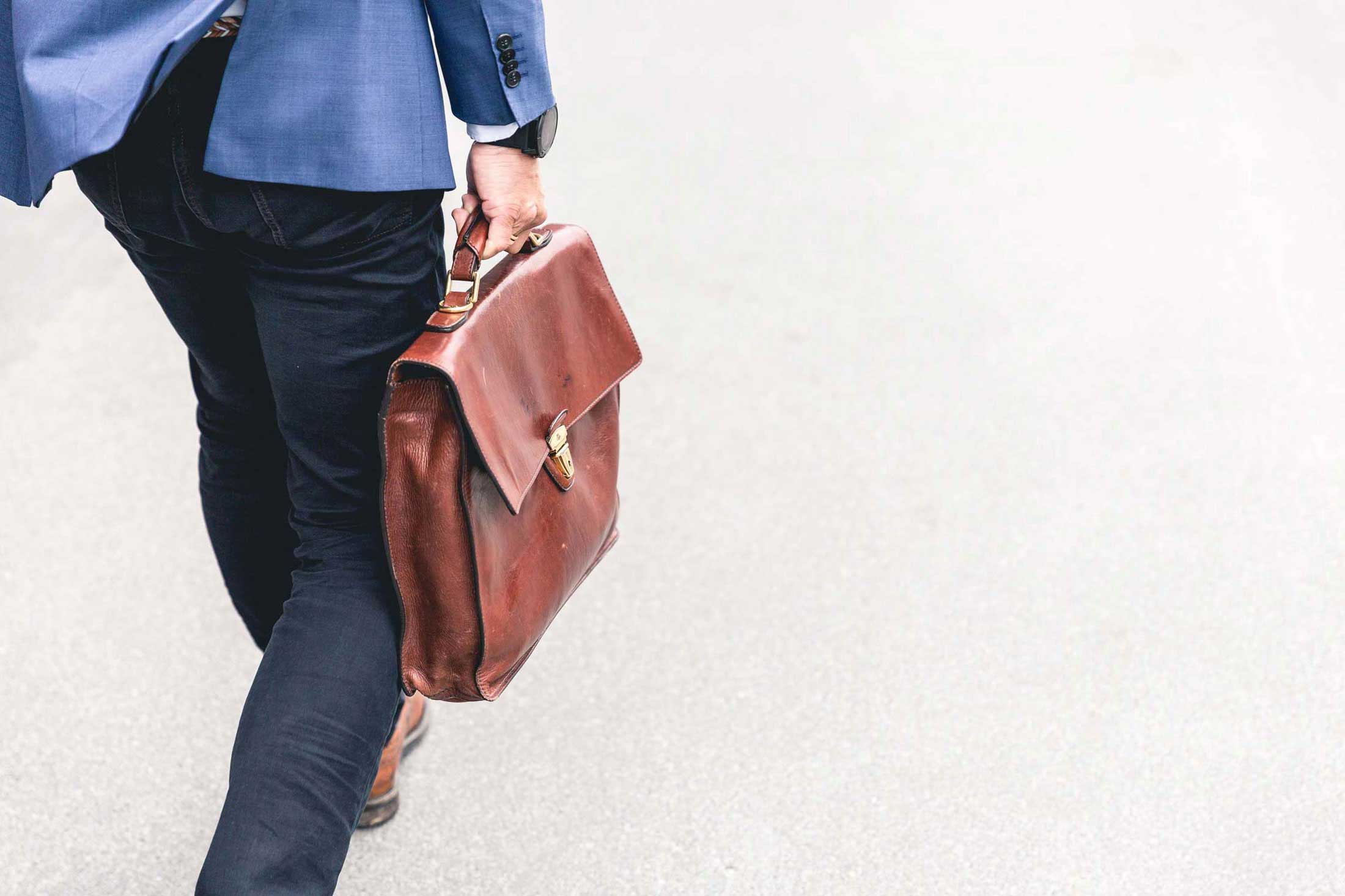 View from the back of a professional man, wearing a suit, walking with a leather briefcase in hand.
